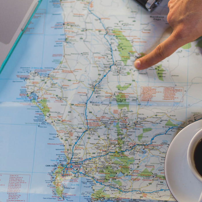 Auf einem Holztisch liegt eine Landkarte, daneben steht eine Tasse mit schwarzem Kaffee und ein Notebook und ein Finger zeigt auf einen Ort auf der Landkarte