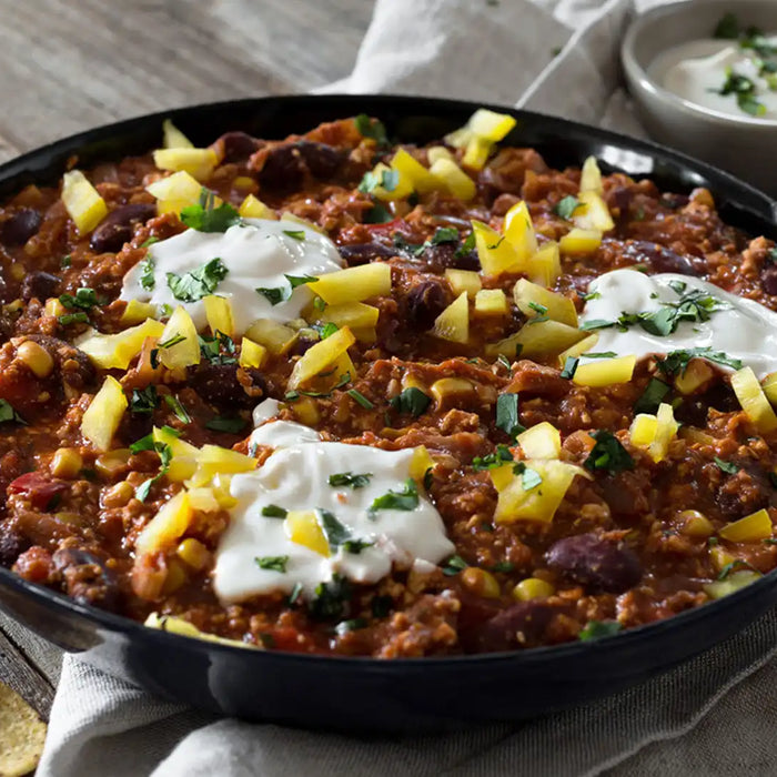 Veganes Chilli mit Kaffee in einer Pfanne auf beigen Leintüchern stehend, ein Kochlöffel, eine Sauce und Tortillas liegen daneben 