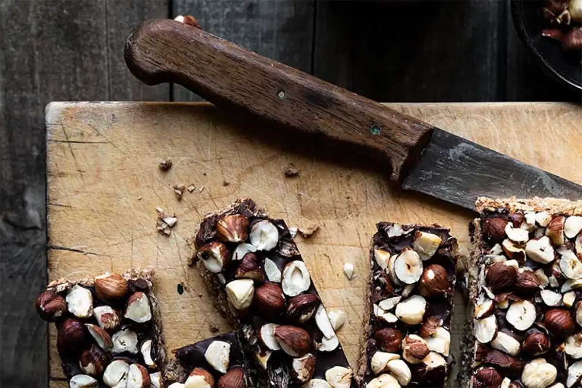 In Stücke geschnittene Coffee-Oat-Bars auf einem Brett mit einem Messer, zerkleinerten Haselnüssen und einer Tasse Kaffee von oben 