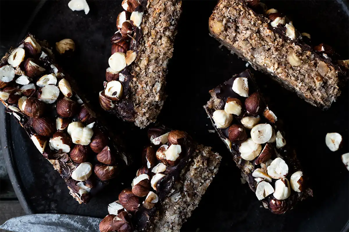 In Stücke geschnittene Coffee-Oat-Bars auf einem dunklen Teller, daneben ein Messer und eine Tasse Kaffee 