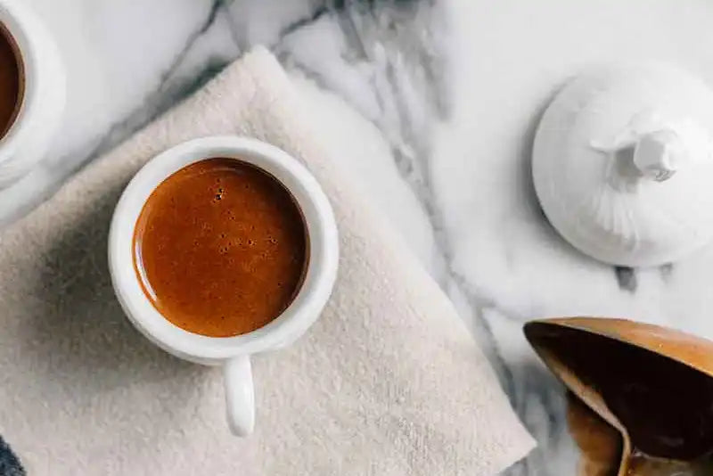 Ein Espresso steht auf einer marmorierten Oberfläche daneben liegt ein Löffel und eine Keramikdose