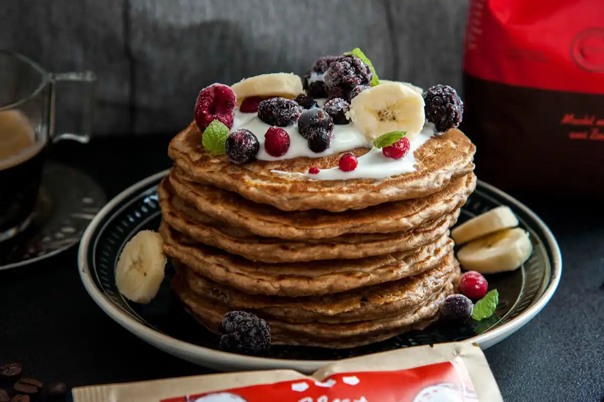 Haferflocken Kaffee Pancakes mit Früchten als Topping auf einem dunklen Untergrund, im Hintergrund steht eine Packung J. Hornig Spezial und eine Tasse Espresso 