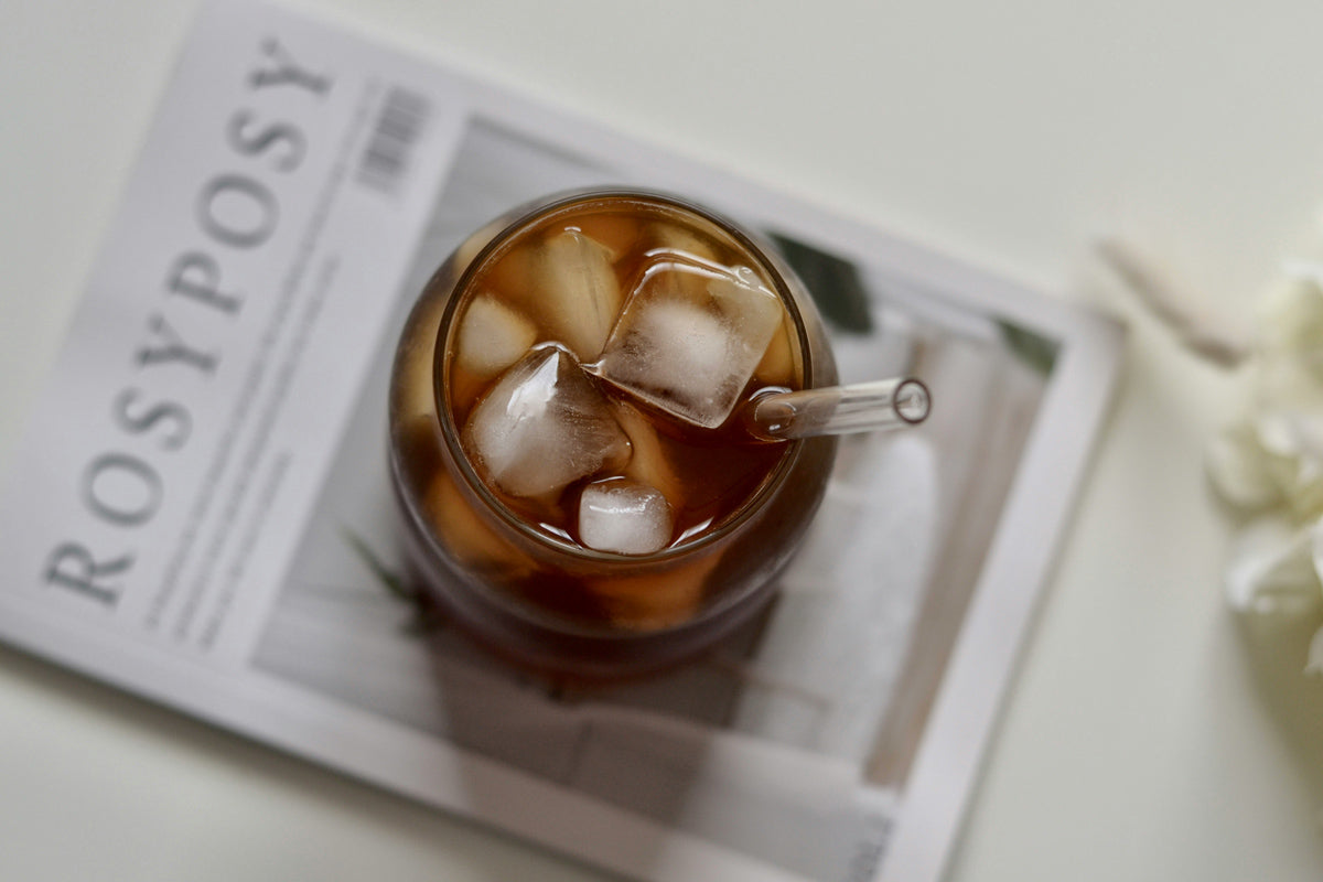 Ein Glas mit Eiswürfel und Kaffee steht auf einer Zeitschrift, welche auf einem Tisch liegt.