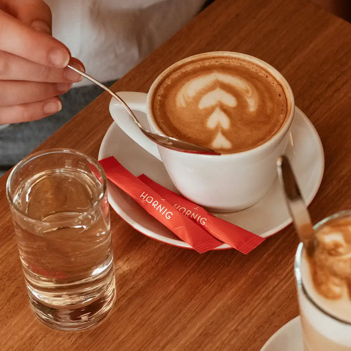 Ein Cappuccino auf einem Holztisch mit zwei roten J. Hornig Zuckersticks.