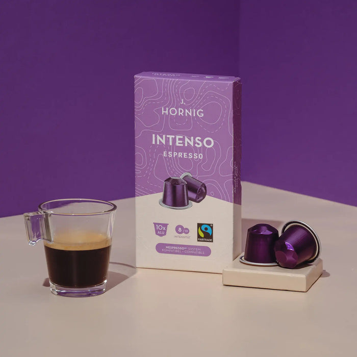 Eine Packung J. Hornig Intenso Espresso Kaffeekapseln mit zwei Alukapseln und einer Kaffeetasse auf hellem Untergrund mit einem grauen Hintergrund