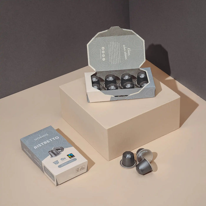 Eine offene Packung J. Hornig Ristretto Kaffeekapseln und eine geschlossen Packung J. Hornig Ristretto Kaffeekapseln auf einem hellen Untergrund mit einem grauen Hintergrund.