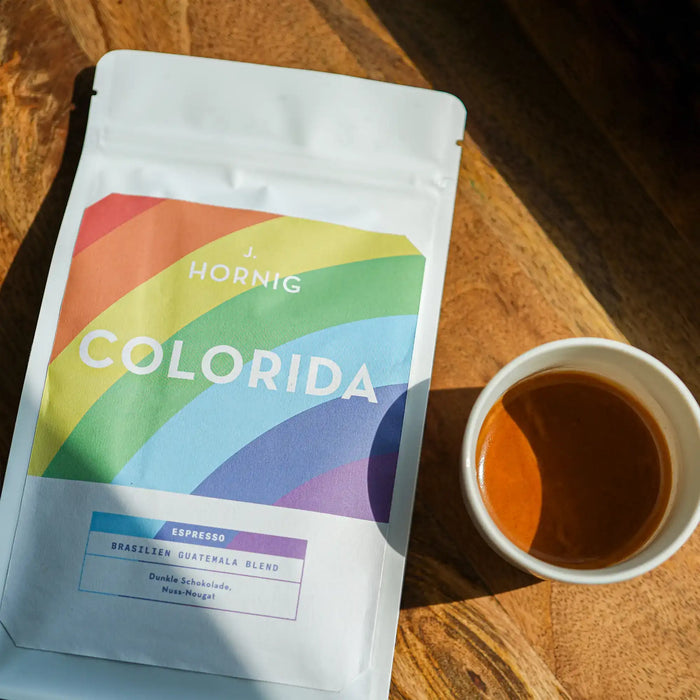 Eine Packung J. Hornig Spezialitätenkaffee Colorida Espresso Limited Edition Brasilien Guatemala Blend liegend auf einem Holztisch mit einer Tasse Espresso