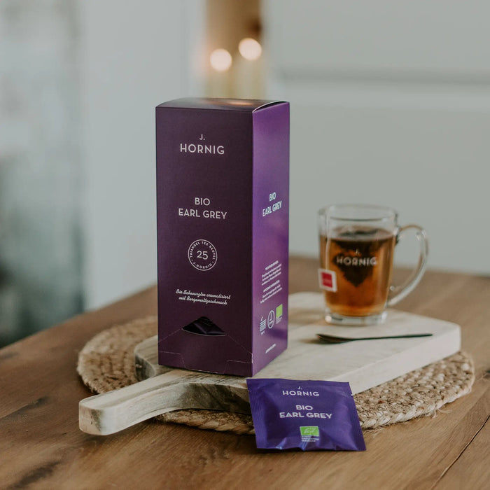 Eine Packung J.Hornig Bio Pyramidenbeutel Earl Grey Tee auf einem Holztisch mit einer Tasse Tee.