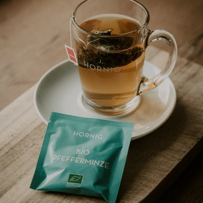Eine Tasse Tee in einem J.Hornig Teeglas mit einer Einzelpackung Bio Pfefferminze Tee.