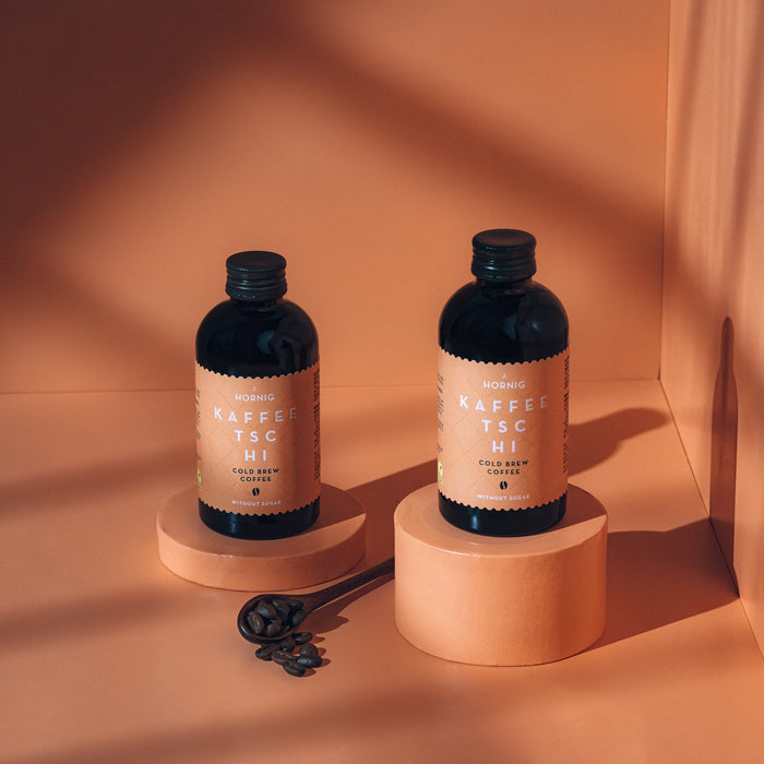 Zwei Flaschen J. Hornig Kaffeetschi Cold Brew auf einem Podest mit einem orangen Hintergrund.