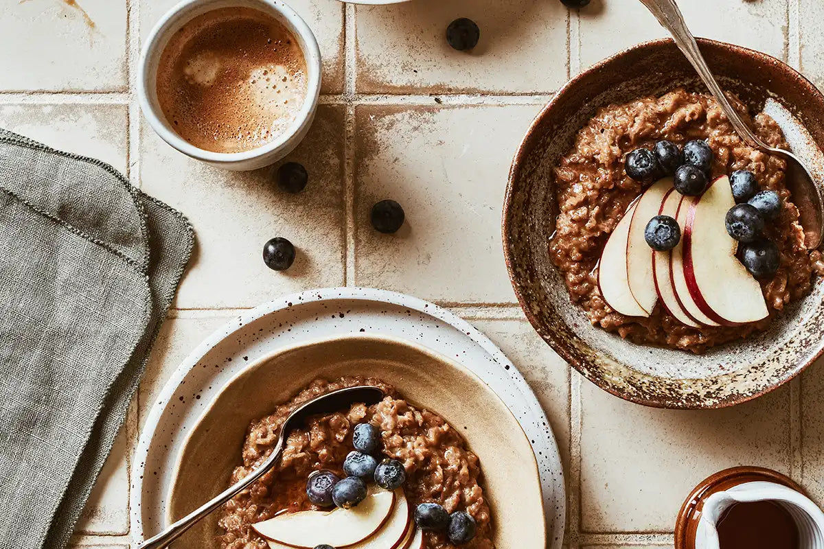  2 Schüsseln mit Kaffee Porridge, garniert mit Heidelbeeren und Äpfeln, und eine Tasse Kaffee stehen auf einem beigen Untergrund 