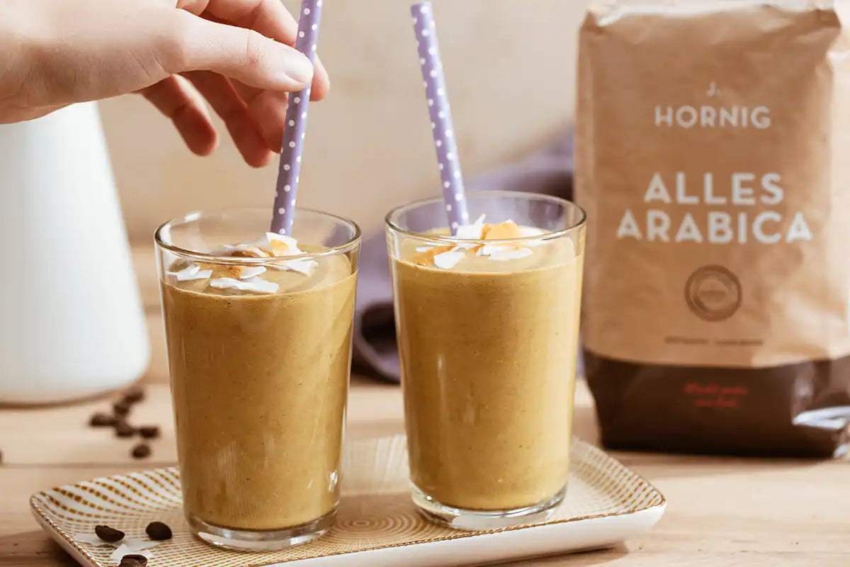 2 Gläser Kaffee Smoothies mit Kokos und Kurkuma stehen auf einem Tablett, eine Hand berührt den Strohhalm und dahinter ist ein Blumenstrauß und der J. Hornig Alles Arabica zu sehen 