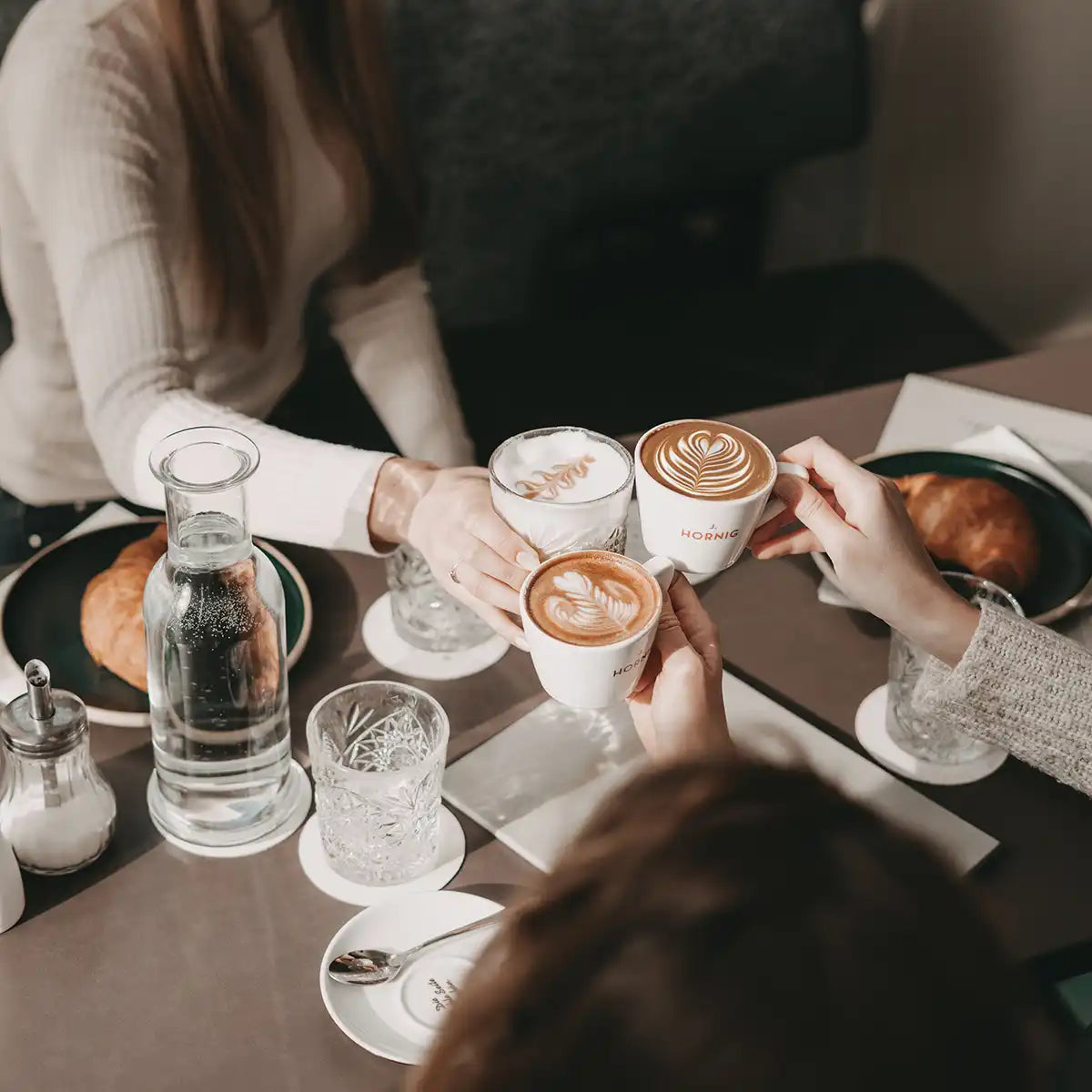 Drei Personen stoßen in einem gemütlichen Kaffeehaus mit ihren Kaffeetassen an.