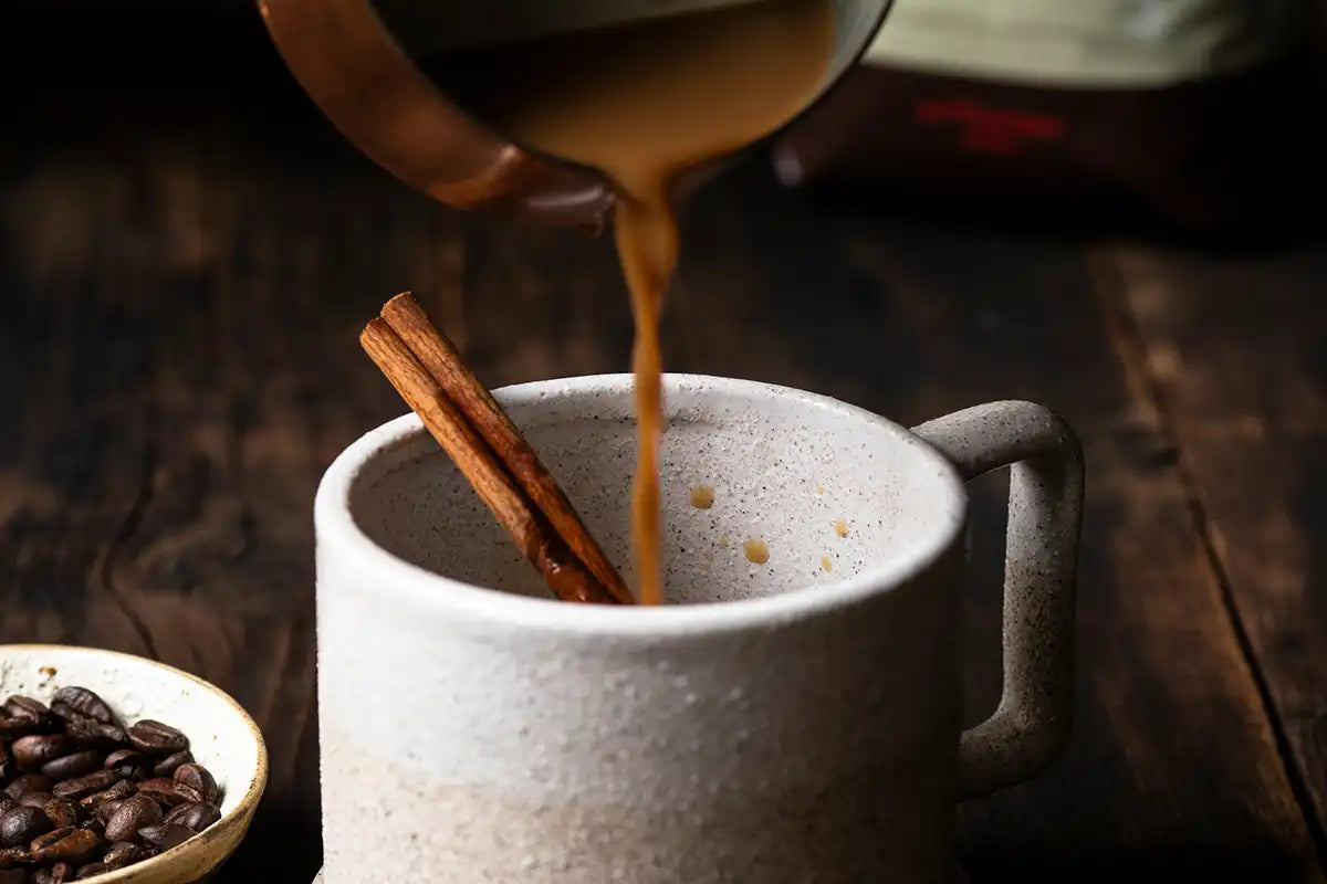 Eine Person schüttet in eine Tasse mit Zimtstange die Kardamom-Zimt-Latte