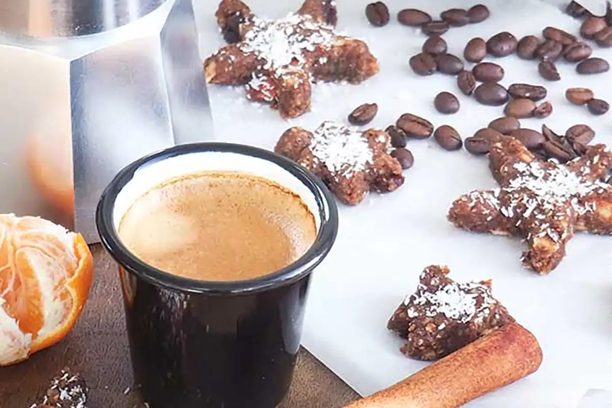 Vegane Kaffee-Weihnachtskekse liegen auf einem Backpapier mit Zimtstangen, einer geschälten Mandarine und daneben steht ein Espressokocher und ein Espresso in einer schwarzen Tasse