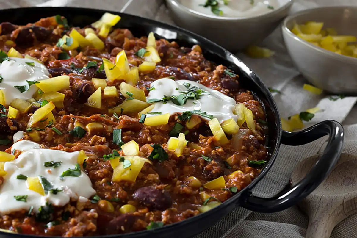 Veganes Chilli mit Kaffee in einer Pfanne auf beigen Leintüchern stehend, ein Kochlöffel und eine Sauce stehen daneben 