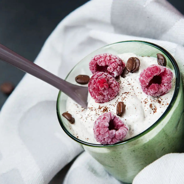 Ein Glas cremiges Kaffee Joghurt mit gefrorenen Himbeeren und Kaffeebohnen auf einem weißen Tuch stehend 