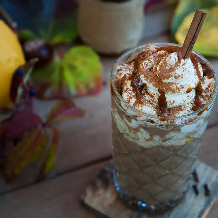 Ein Glas mit Pumpkin-Spice-Latte verziert mit Schlag, Zimt und Zimtstande steht neben bunten Blättern und einem gelben Kürbis 