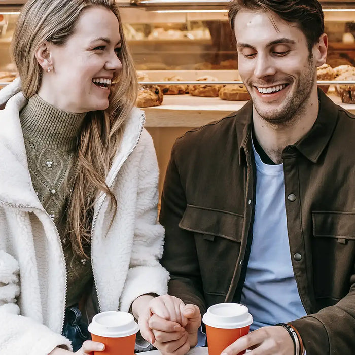 Ein Paar hält vor einem Coffeeshop Händchen, lacht sich an und hält in der anderen Hand rote Coffee-To-Go Becher