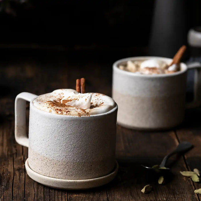 Zwei Becher mit Kardamom-Zimt-Latte garniert mit einer Zimtstange stehen auf einem Holzbrett 