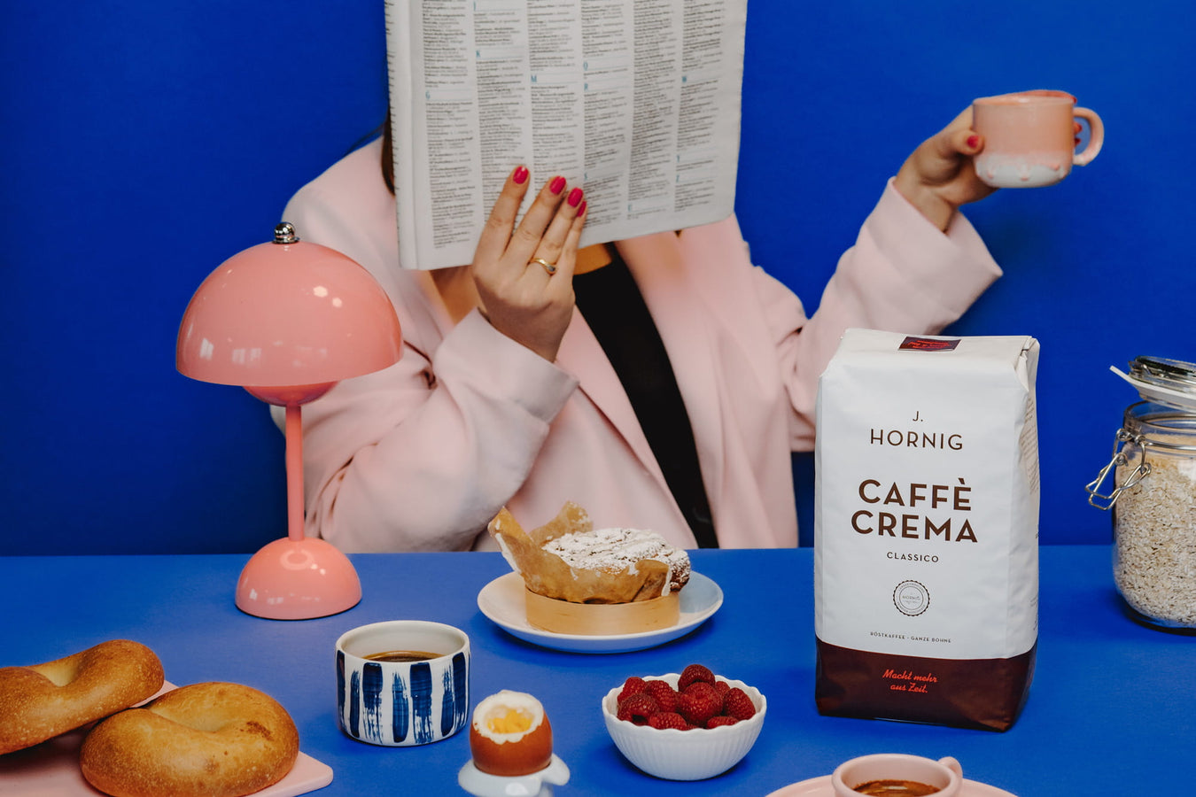 J. Hornig Caffè Crema Classico auf Frühstückstisch mit Croissants, Beeren und Espressotasse, Person liest Zeitung im Hintergrund