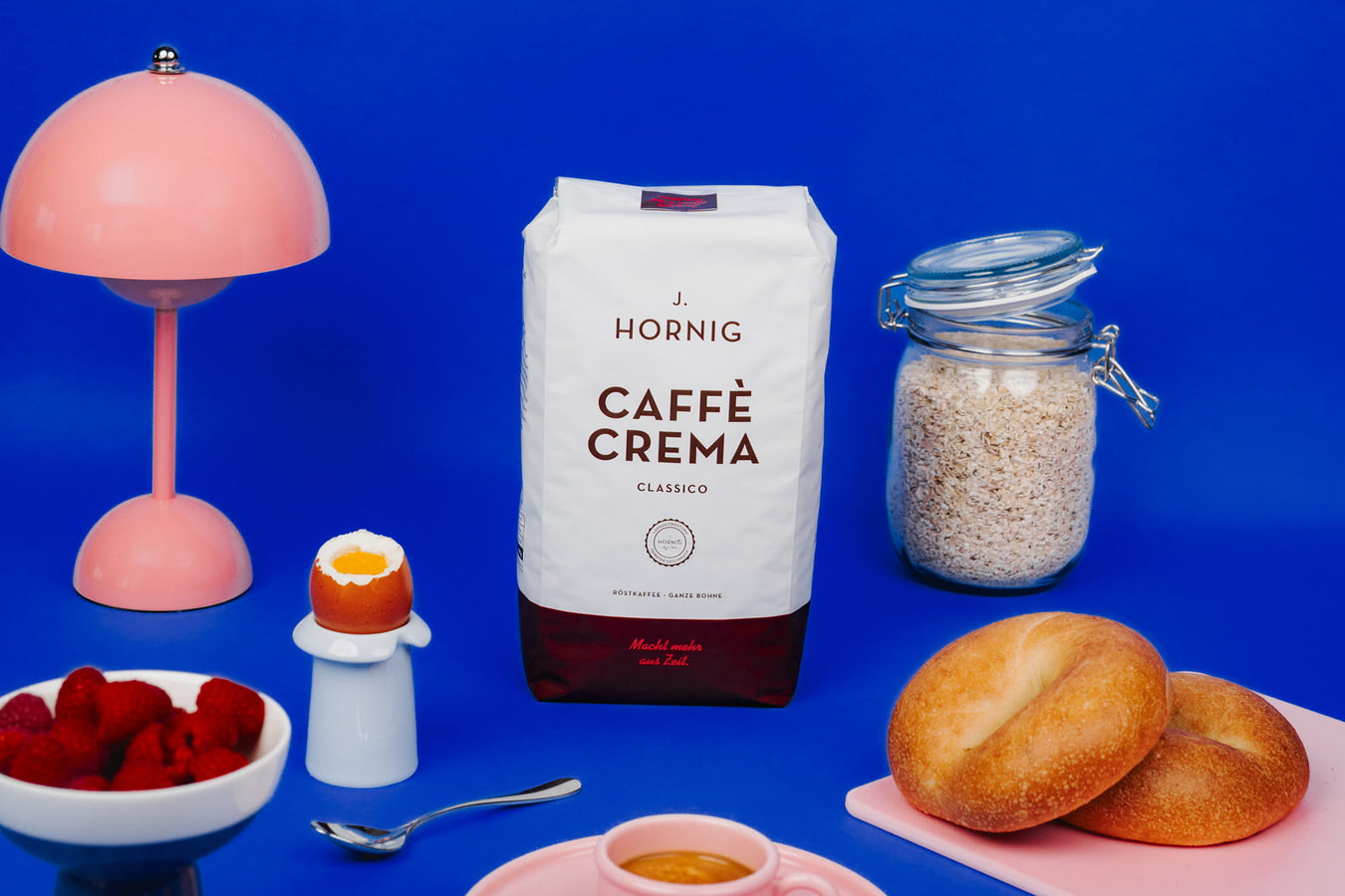 J. Hornig Caffè Crema Classico mit Haferflocken, Beeren, Brot und Espresso in modernem Frühstücks-Arrangement vor blauem Hintergrund