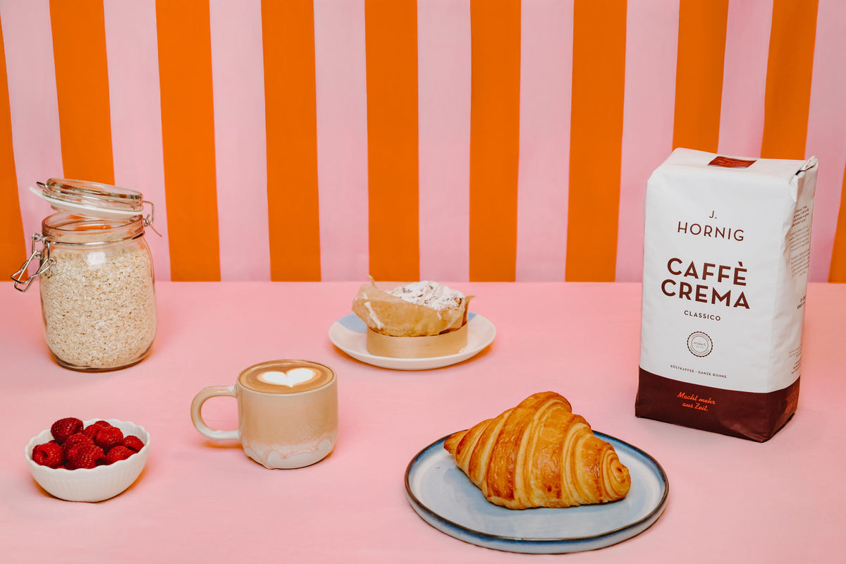 J. Hornig Caffè Crema Classico inszeniert mit Croissant, Cappuccino und Müsli auf rosa Fläche mit orangefarbenem Streifenhintergrund