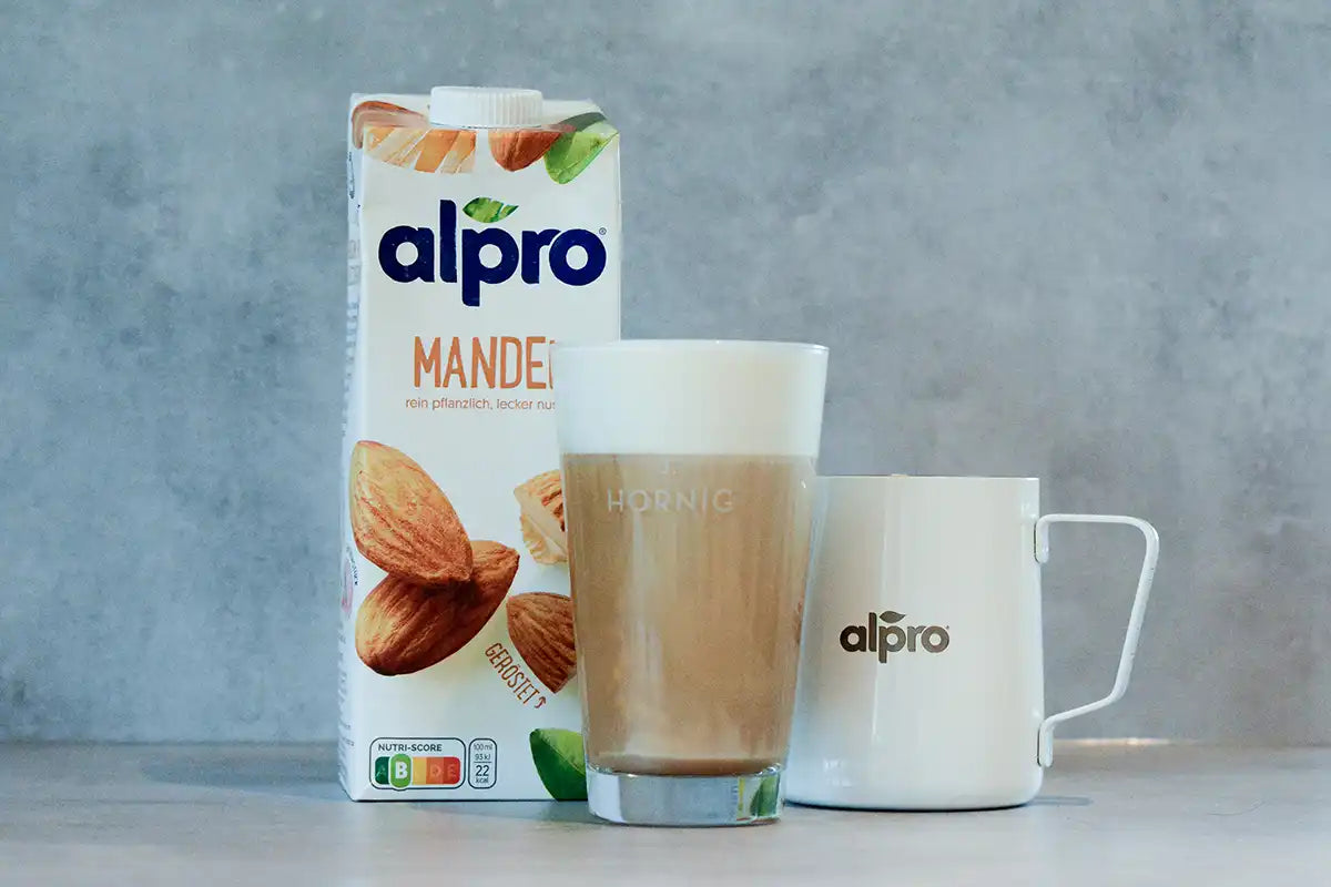 Eine Packung Alpro Mandel steht neben einem Caffe Latte und einer Alpro Milchkanne