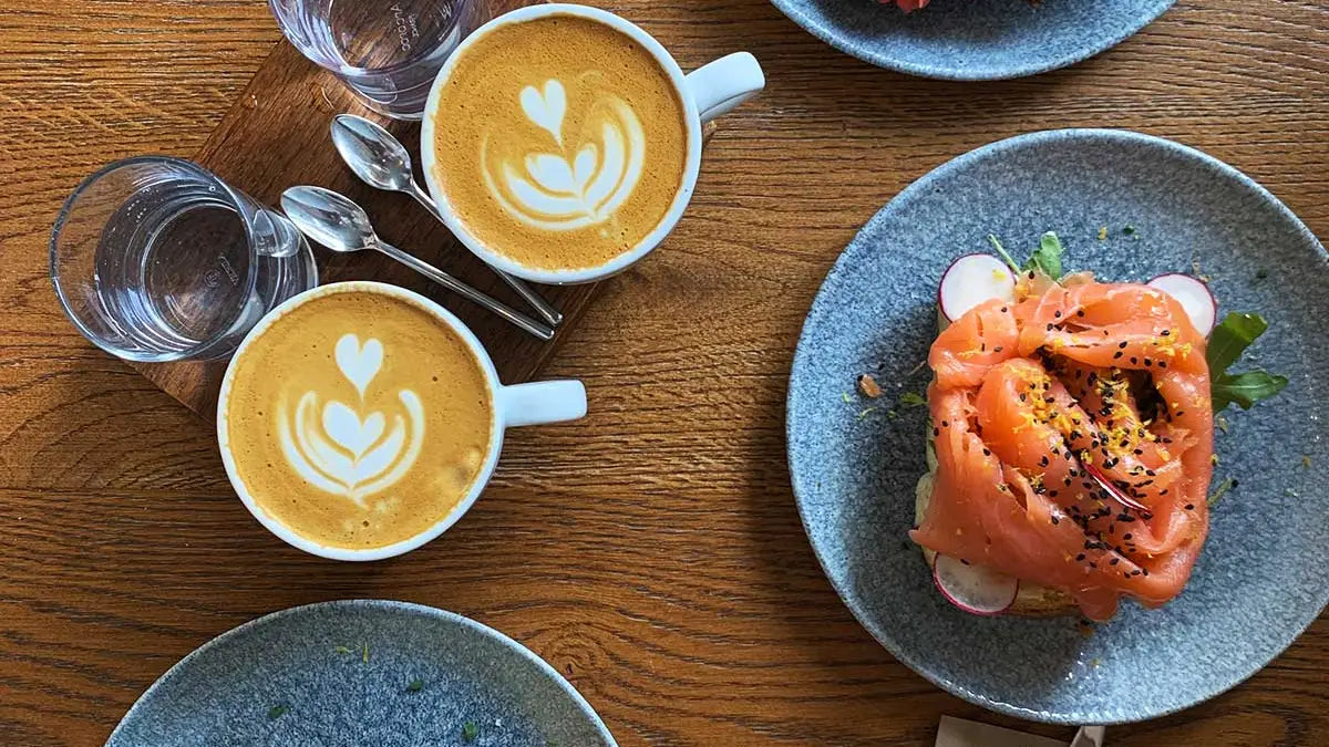 : 2 Cappuccino mit Wassergläser stehen auf einem Holztisch daneben steht ein Teller mit einem Lachsbrötchen