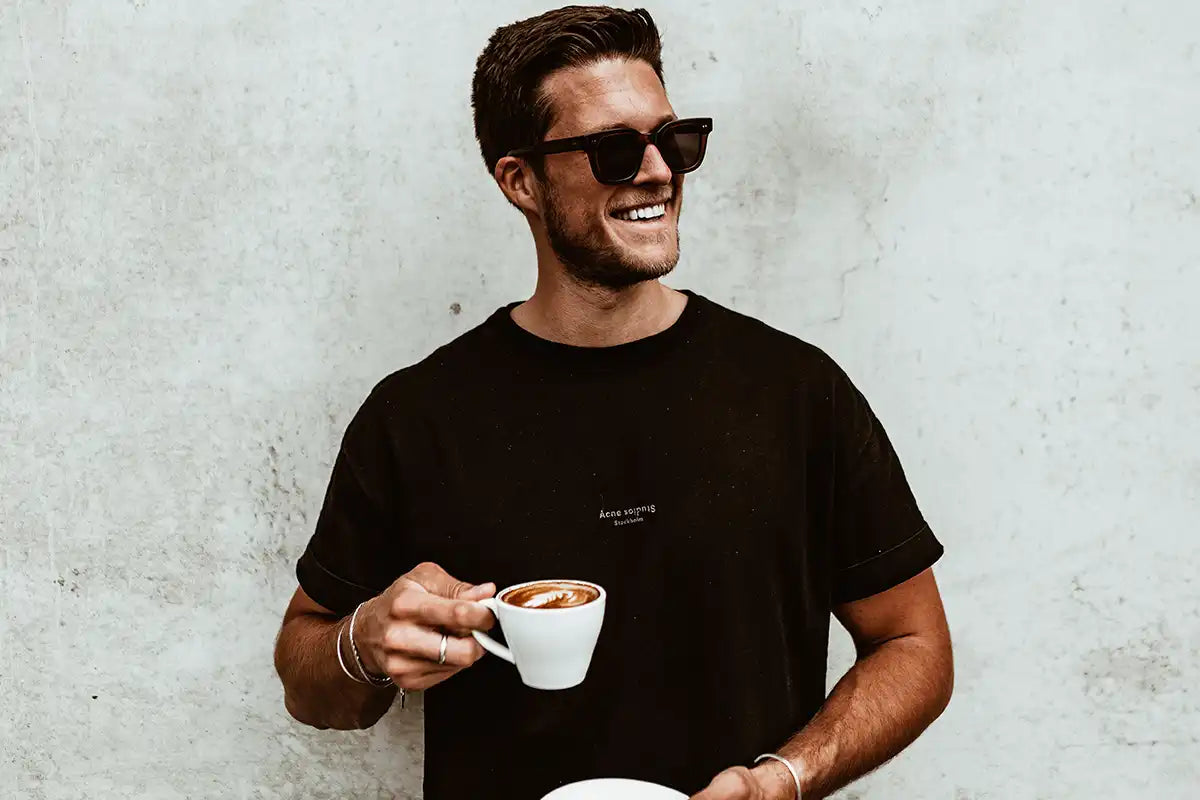 Eine junge Person mit einer schwarzen Sonnenbrille hält eine Tasse Kaffee in der Hand.