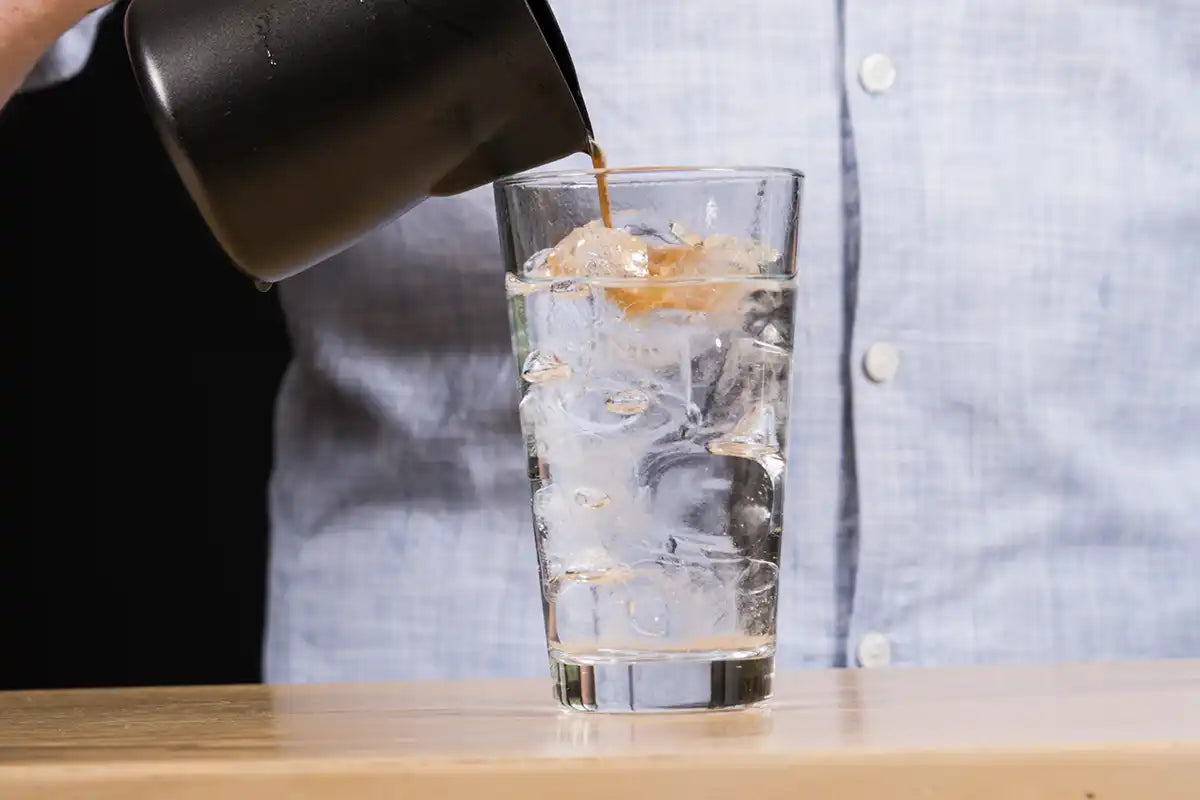 Ein Mann schüttet mit einer Hand Kaffee in ein Glas mit Eiswürfeln und Tonic