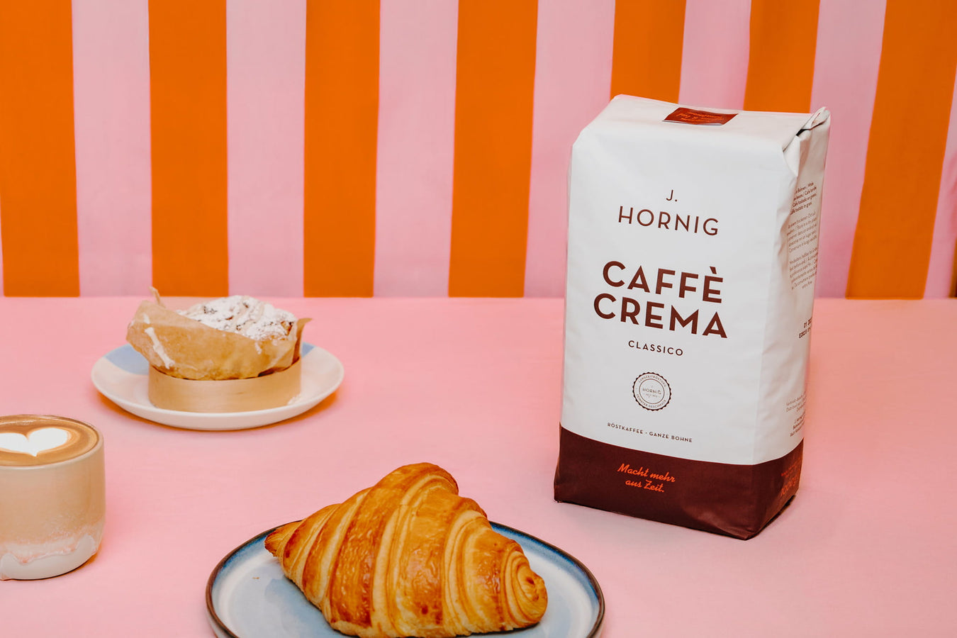 J. Hornig Caffè Crema Classico mit Croissant, Cappuccino und Frühstücksgedeck auf rosafarbenem Tisch vor gestreiftem Hintergrund