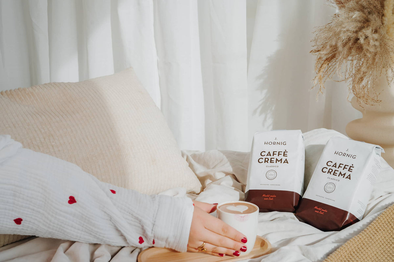 Eine Tasse Kaffee mit zwei J. Hornig Caffe Crema Classico Packungen auf hellem Bett mit Vorhängen im Hintergrund.