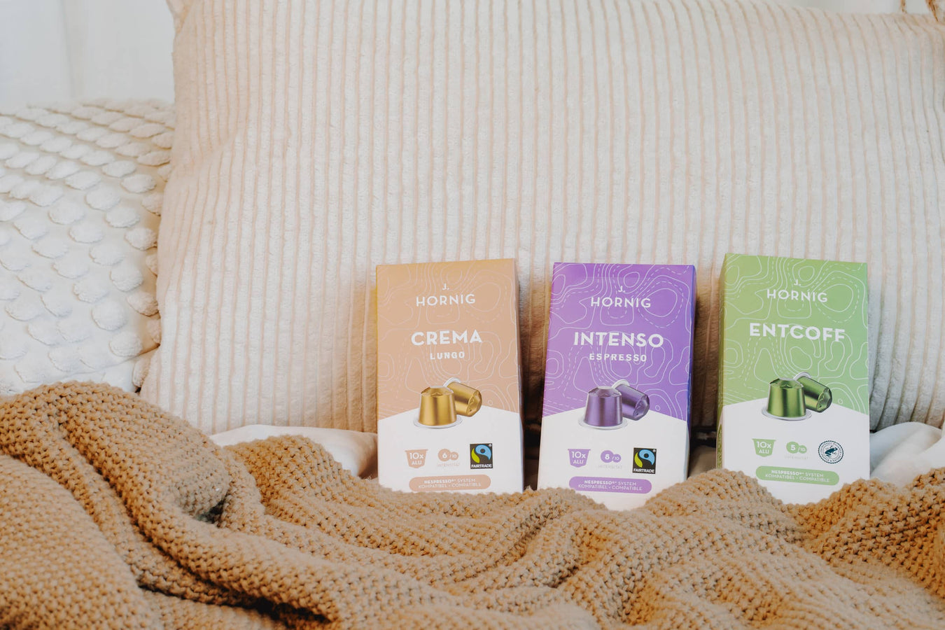 Drei Packungen J. Hornig Kaffeekapseln – Crema Lungo, Intenso Espresso und Entcoff – auf einer beigen Decke neben einer Tasse Kaffee und Kissen.
