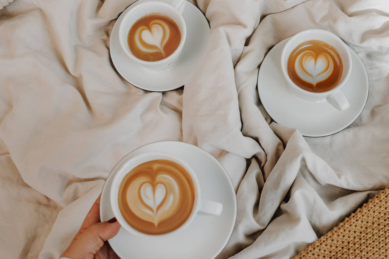 Drei Cappuccinos mit Latte Art auf weiß-beiger Bettdecke.
