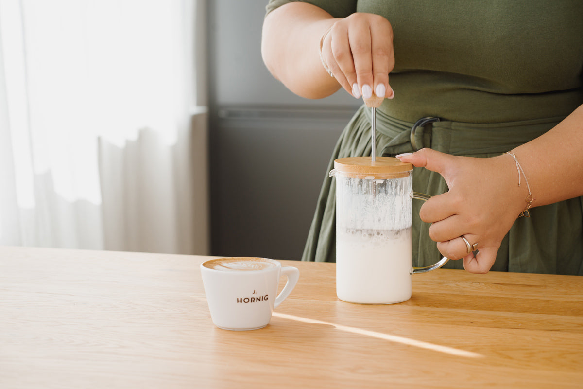 Milch ist in einer Frenchpress, daneben steht ein Cappuccino.