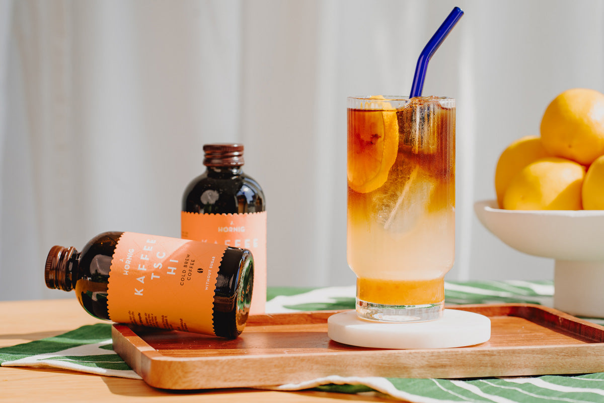 Fertiger Yuzu Cold Brew Latte steht auf einem Tisch. Daneben zwei Flaschen J. Hornig Kaffeetschi Cold Brew. 