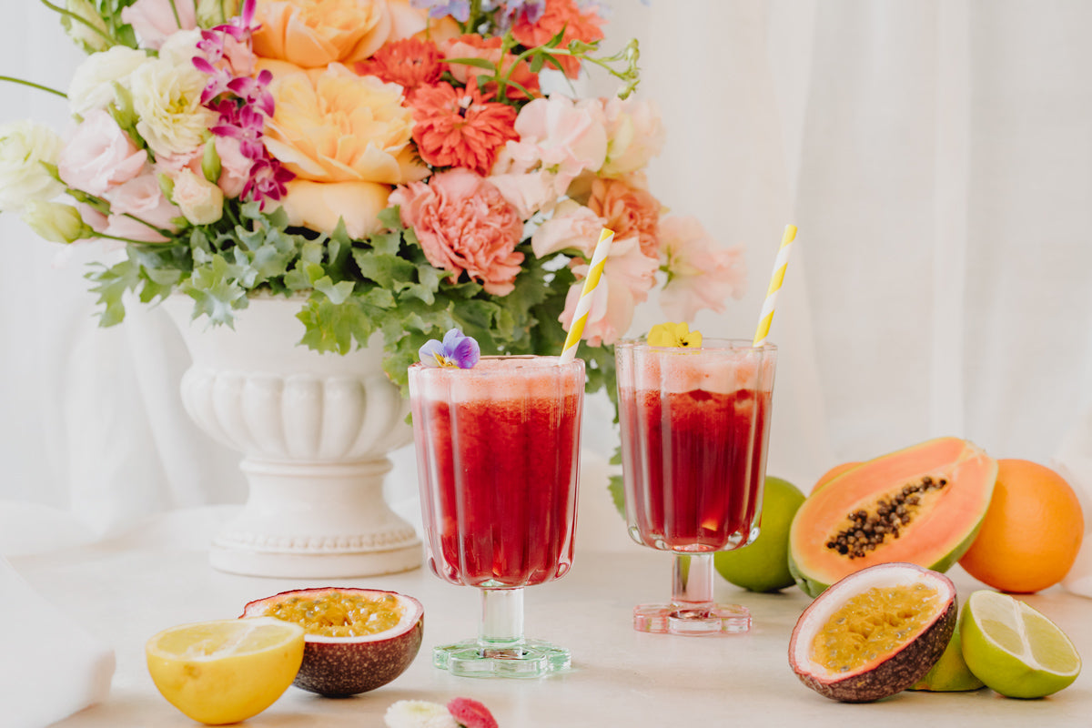 Zwei Cocktails stehen auf einem sommerlich dekorierten Tablett mit Blumen im Hintergrund. 