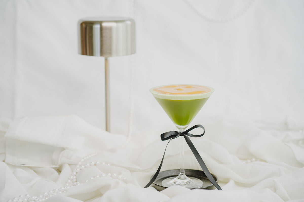 Matcha Martini mit Espresso Foam mit Schleife am Glas mit Perlen und einer Lampe im Hintergrund.