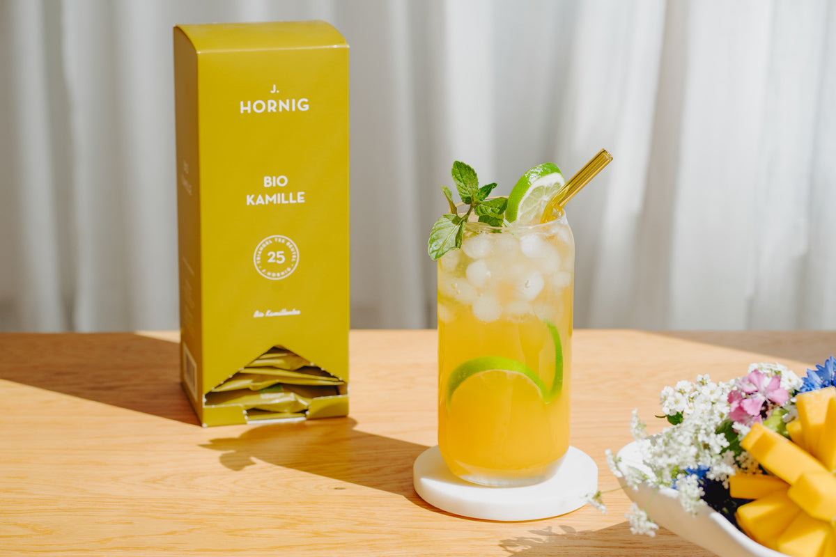 Der Eistee steht auf einem Tisch, im Hintergrund sind Mango, Blumen und Kamillentee zu sehen.