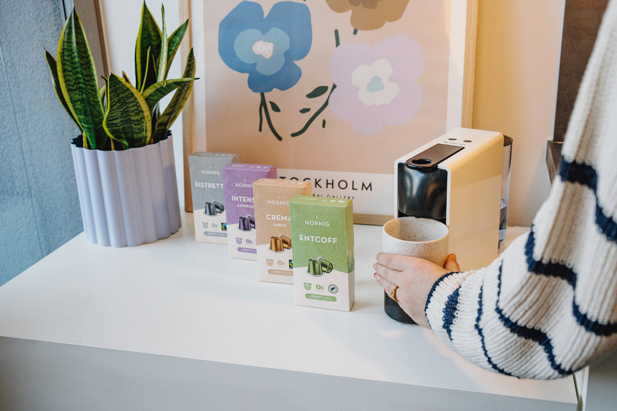 Neben einer Kapselkaffeemaschine liegen mehrere J. Hornig Kaffeekapseln. Eine Hand nimmt eine frisch gebrühte Tasse von der Maschine.