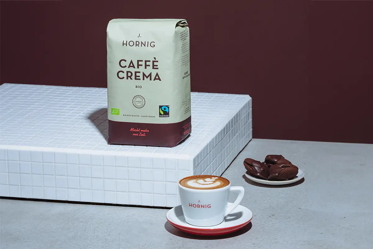 Eine Packung Caffe Crema Bio auf einem weißen Podest mit einer Tasse Kaffee und Gebäck.