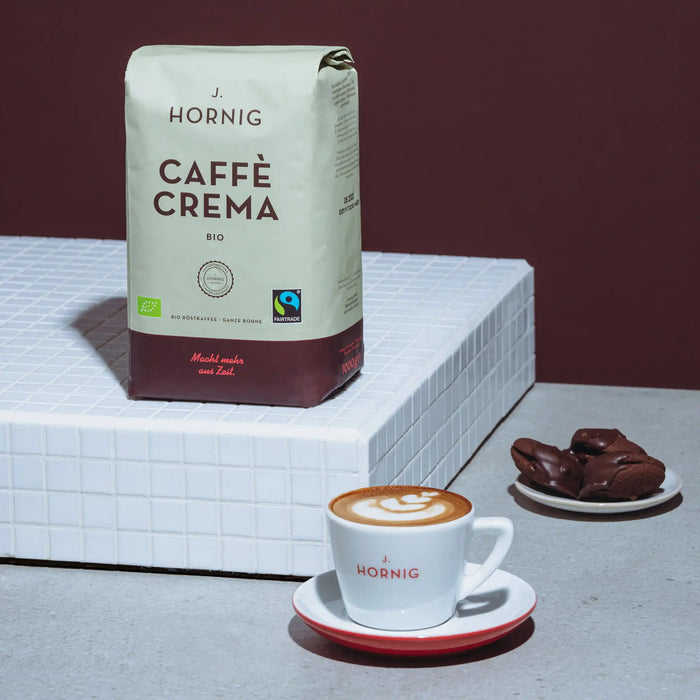 Eine Packung J. Hornig Caffe Crema Bio Kaffee mit einer Cappuccinotasse und Gebäck auf einem weissen Podest mit braunem Hintergrund