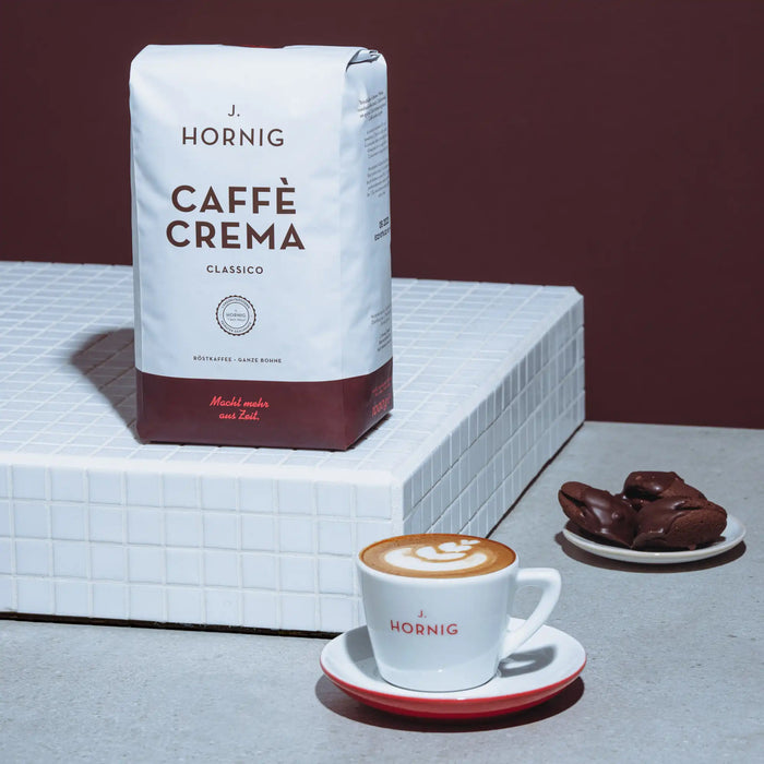 Eine Packung J. Hornig Caffe Crema Classico mit einer Cappuccinotasse und Gebäck auf einem weißen Podest mit braunem Hintergrund