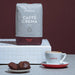 Eine Packung J. Hornig Caffe Crema Intenso auf einem weißen Podest mit einem Cappuccino und Gebäck