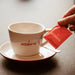 Eine J. Hornig Cappuccino Tasse mit einem rotem J. Hornig Zuckerbriefchen.
