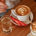 Ein Cappuccino auf einem Holztisch mit zwei roten J. Hornig Zuckersticks.