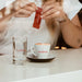 Eine Person öffnet einen J. Hornig Karamellkeks mit einem Cappuccino und einem Glas Wasser.