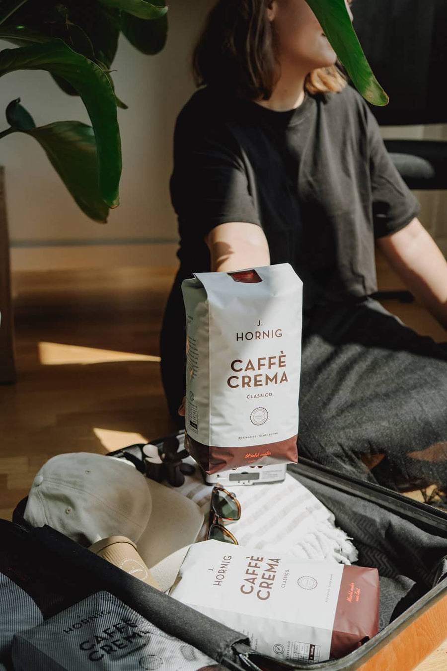 Frau sitzt neben offenem Koffer mit Sonnenbrille, Cap und Handtuch; hält eine Packung J. Hornig Caffè Crema Classico ins Sonnenlicht.