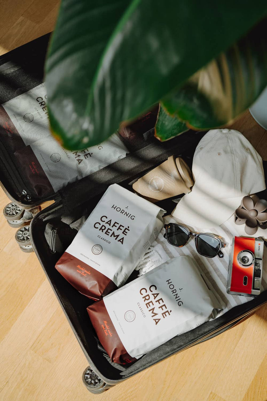 Zwei Packungen J. Hornig Caffè Crema Classico im geöffneten Reisekoffer mit Sonnenbrille, Retro-Kamera, Coffee-to-go-Becher, Cap und Handtuch – bereit für den Urlaub.